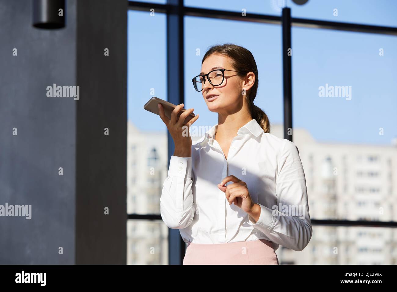 Intelligente junge Geschäftsfrau in stilvoller Brille, die im modernen Büro steht und mit dem Smartphone unterwegs ist, während sie zur Kommunikation Audio-Nachrichten aufzeichnet Stockfoto