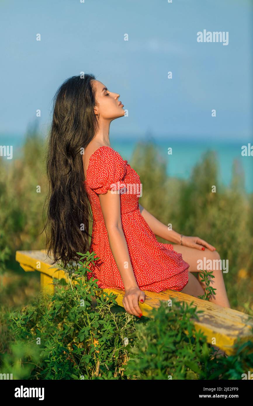 Mädchen in einem roten Kleid mit Tupfen sitzt auf einer Bank mit Blick auf das Meer Stockfoto