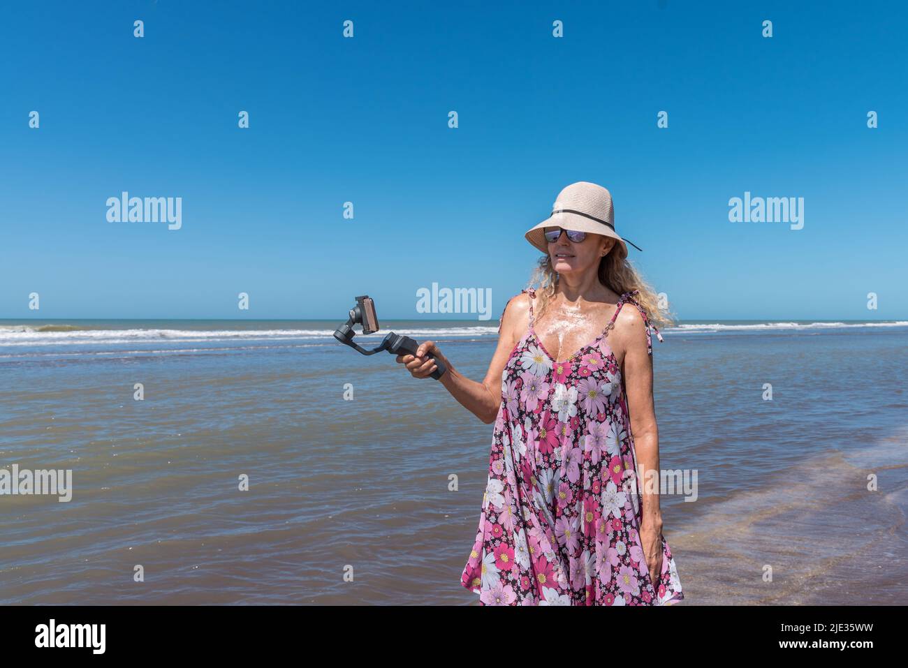 Frau mit einem Hut, der am Strand steht, während sie mit einem auf einem Gimbal-Stabilisator befestigten Handy filmt Stockfoto