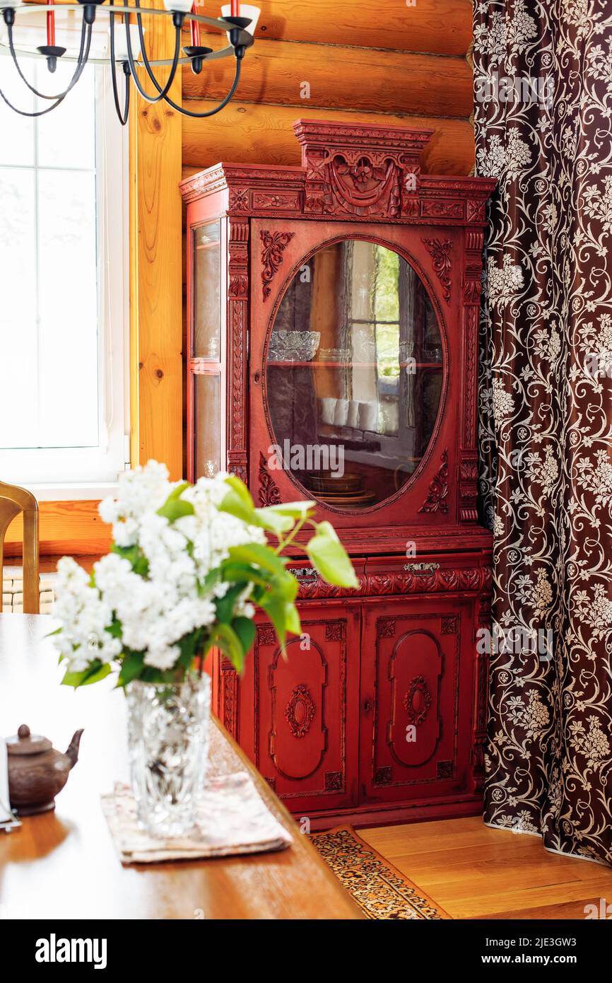 Alter antiker roter Holzschrank mit geschnitzten Dekorationen in der Ecke der Blockhütte in der Nähe eines braunen Vorhangs mit Blumenmustern. Stockfoto