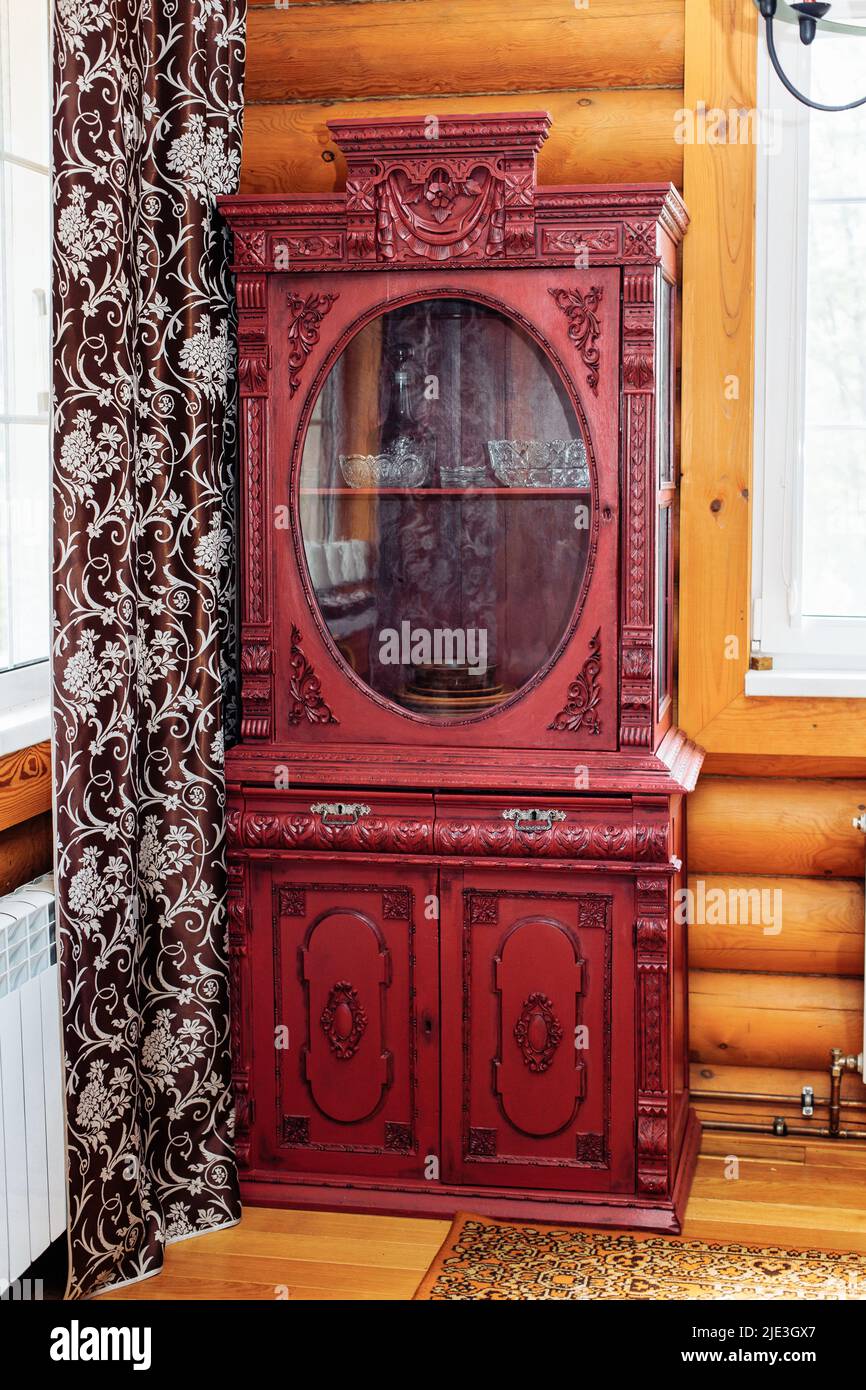 Alter alter, roter Holzschrank mit geschnitzten Dekorationen in der Ecke einer ländlichen Blockhütte zwischen Fenster und braunem Vorhang. Stockfoto