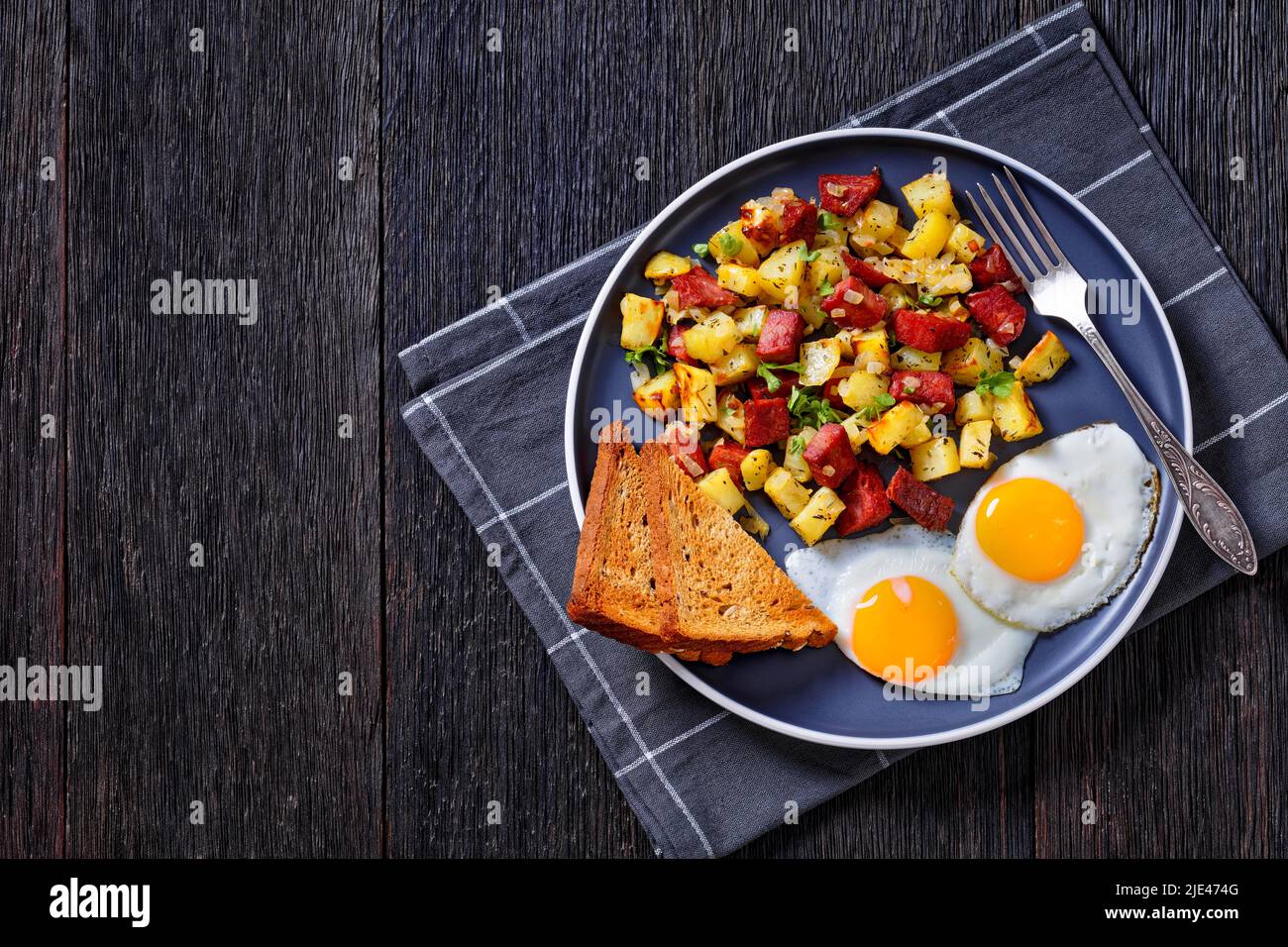 Corned Beef und Hashbrowns, Spiegeleier und geröstetes Roggenbrot auf Teller auf dunklem Holztisch, horizontale Ansicht von oben, flach liegend Stockfoto