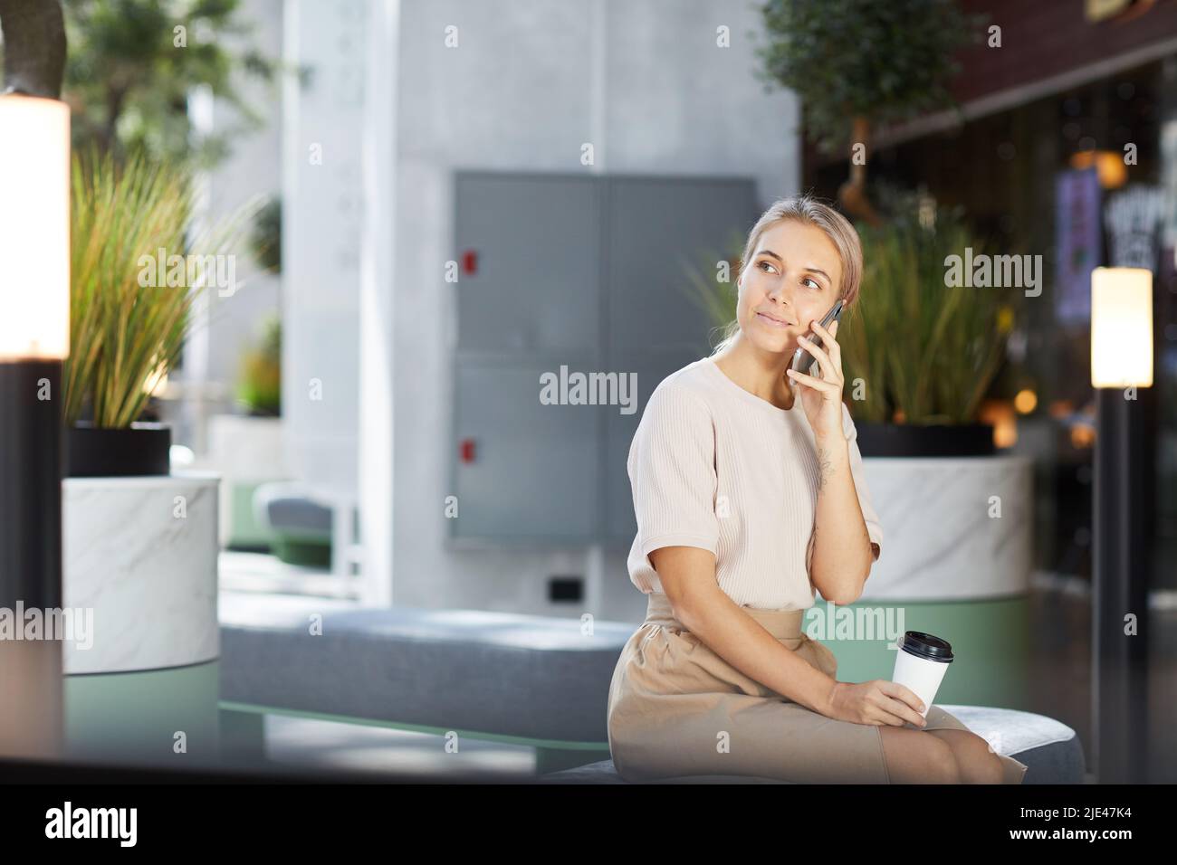 Inhalt hübsche Geschäftsfrau in stilvollem Outfit, sitzt auf einer bequemen Bank in der Lobby und hält einen Takeout-Becher, während sie per Telefon anruft Stockfoto