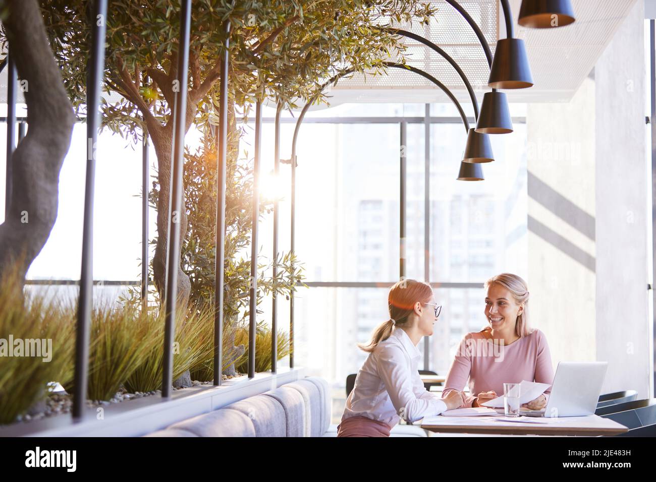 Positive junge unternehmungslustige Geschäftsfrauen sitzen am Tisch im Restaurant mit Pflanzen und diskutieren Bericht, sie genießen Teamarbeit Stockfoto