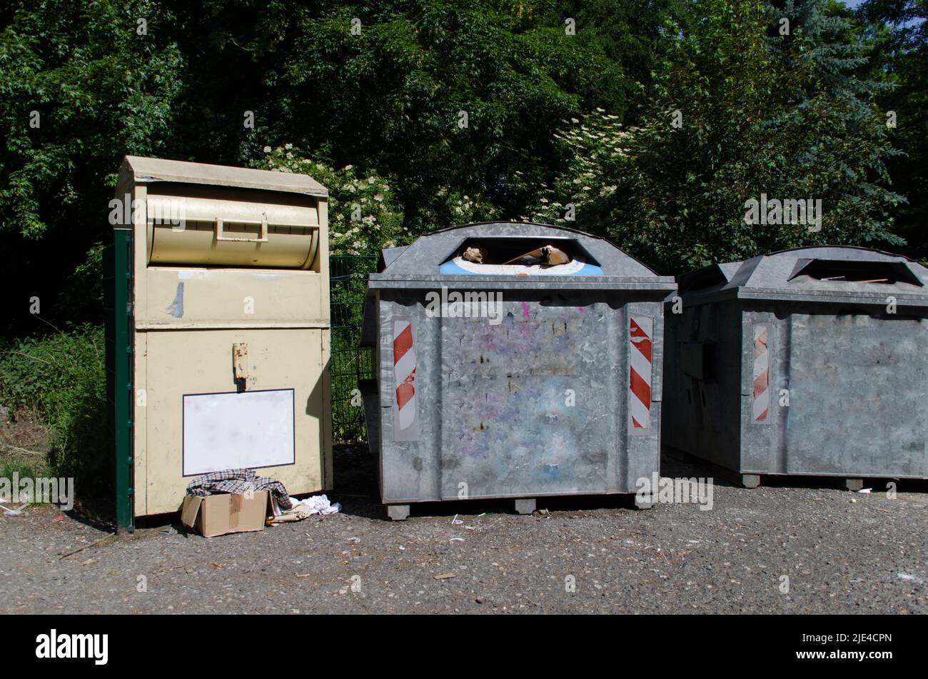 Gruppe von einem Kleidersammelbehälter mit Kleidung auf dem Boden und zwei vollen Papierabfällen in einem Recyclingzentrum oder einer Sammelstation Stockfoto