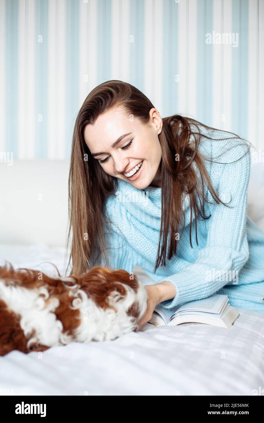Junge hübsche Frau mit langen Haaren trägt blauen Strickpullover auf dem Bett liegend mit Hund im Schlafzimmer, Tierbesitzer Stockfoto