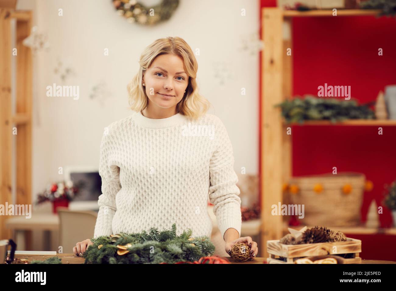 Portrait of content schönes Mädchen in weißen Pullover stehen in gemütlichen Handwerk Studio und machen Weihnachtskranz Stockfoto