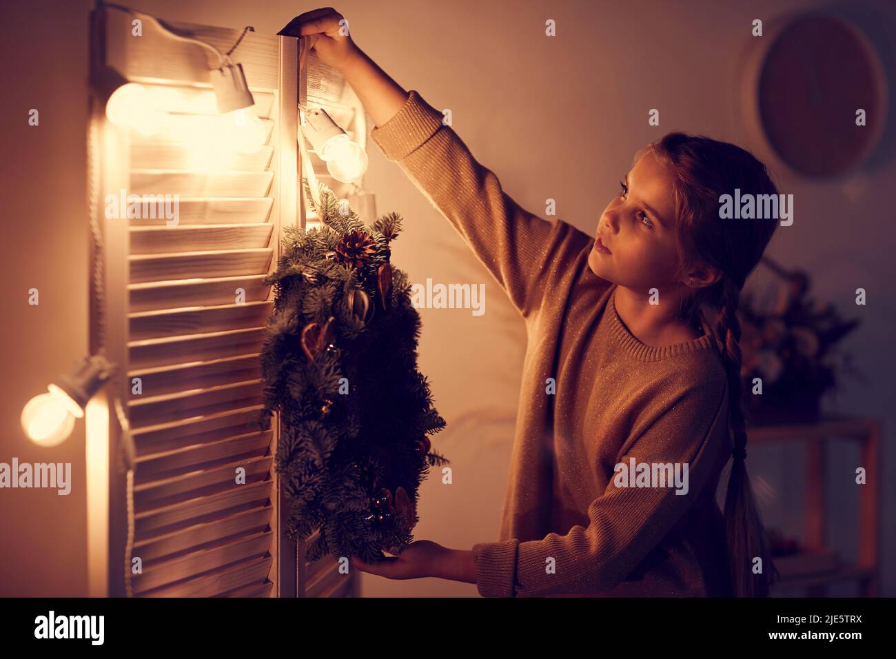 Nettes Mädchen mit Zöpfen tragen beige Pullover stehen am Klappbildschirm und hängen Urlaub Kranz während der Vorbereitung eigenen Raum für Weihnachten Stockfoto