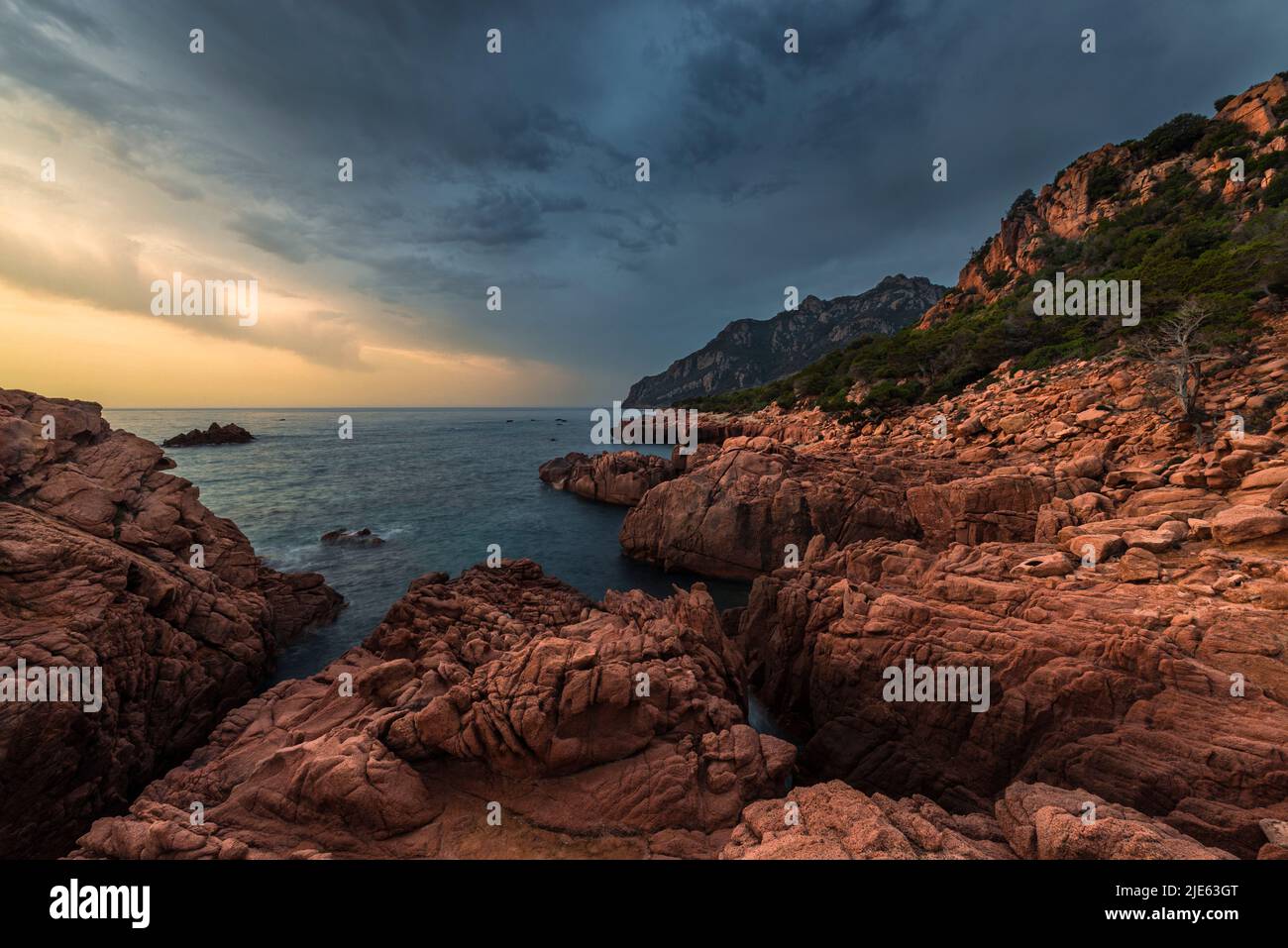 Sonnenaufgang mit dramatischem Himmel an der Bucht von Coccorocci mit den roten Porphyrfelsen, grauen Kieselsteinen und Monte Cartucceddu an der Ostküste Sardiniens Stockfoto