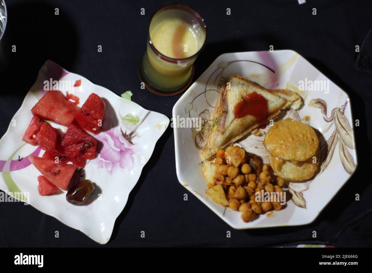 Leckeres Frühstück Orangenmangosaft im Glas. Sandwiches und Snacks auf dem Teller mit frischen Datteln und Wassermelone auf schwarzem Hintergrund Stockfoto