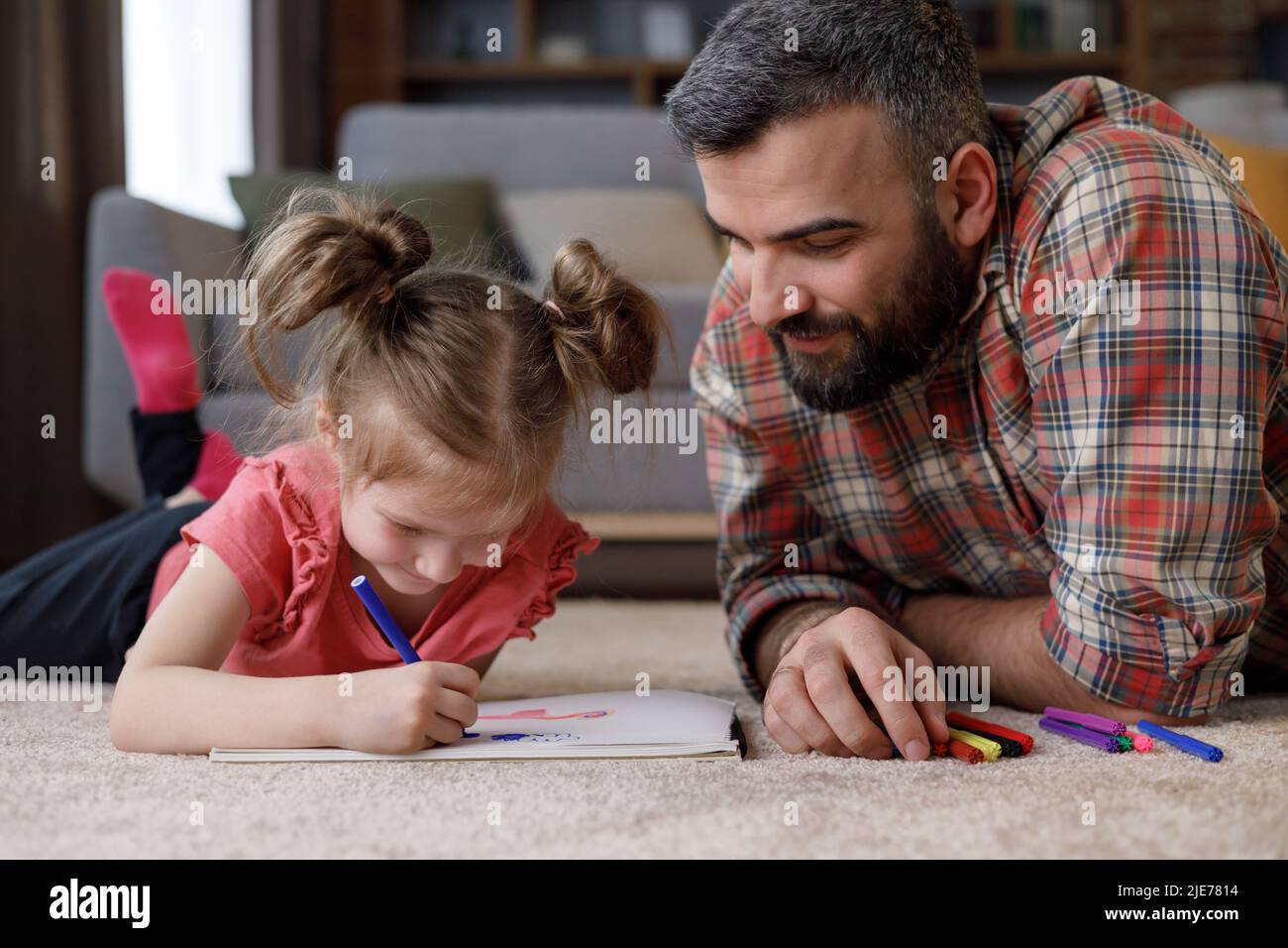 Junger, hübscher Vater und niedliche Tochter spielen zusammen. Vater und Kind malen Bilder, die auf dem Teppich in der Nähe des Sofas zu Hause liegen. Viel Spaß mit Ausgaben Stockfoto