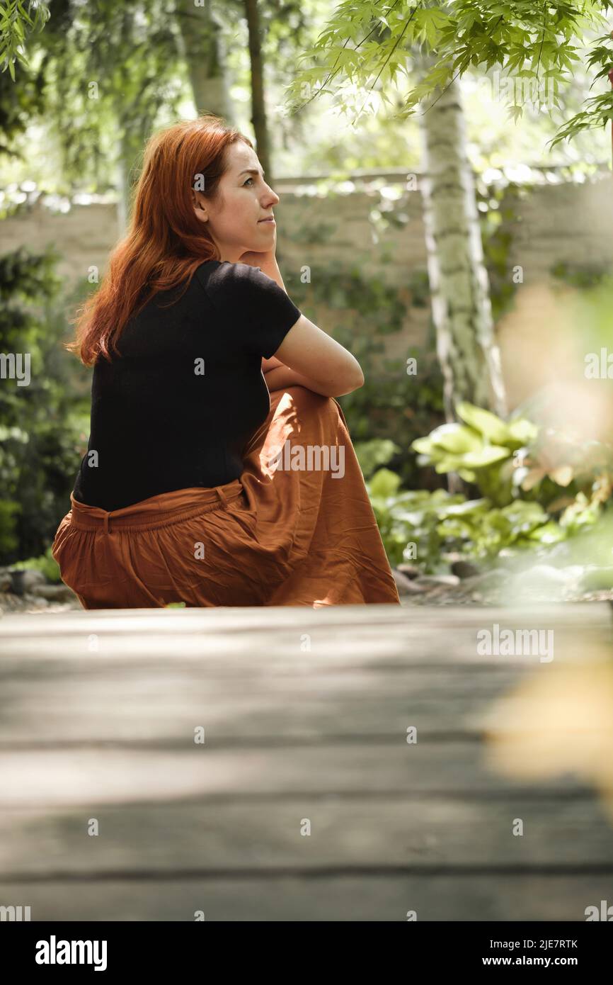 Frau mit 40s Jahren sitzt auf dem Boden und blickt weg in einen grünen Garten. Ruhe haben, denken. Weibchen mit Ingwerhaar von hinten auf der Brücke Stockfoto