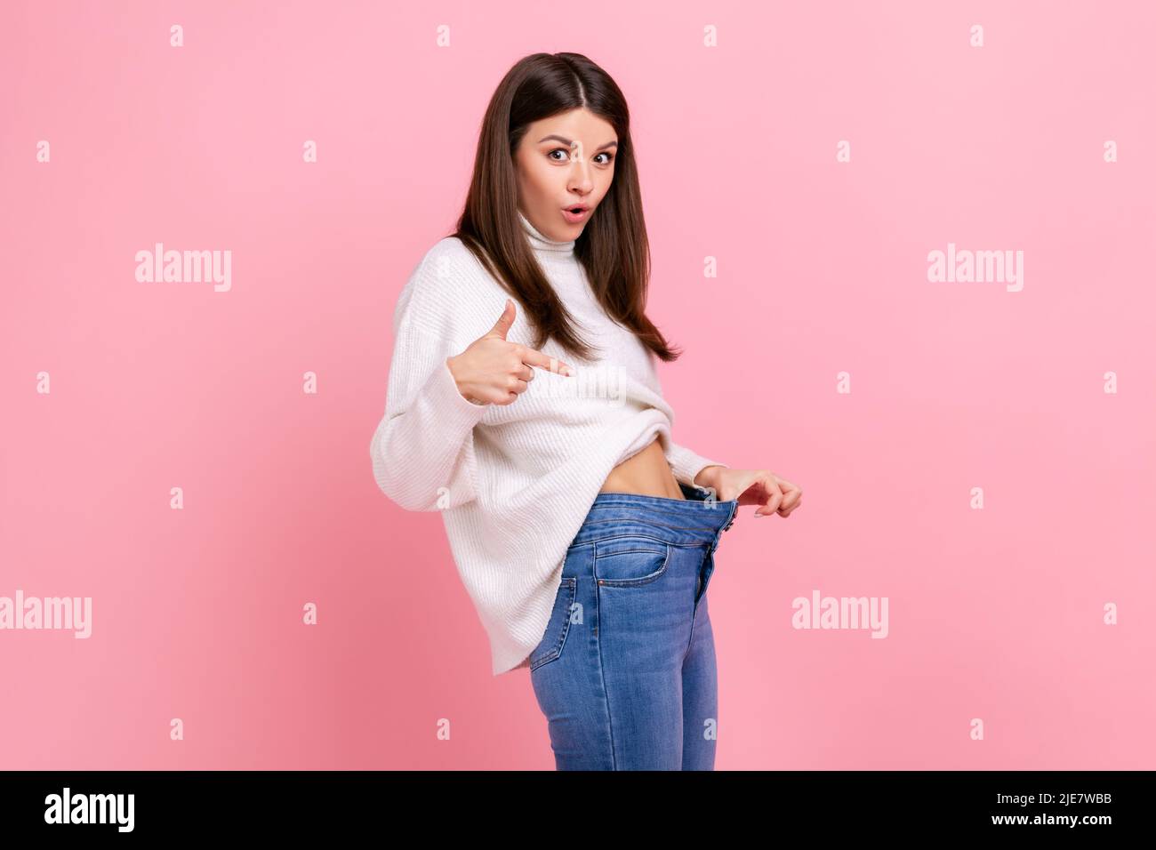 Portrait der schönen überrascht Mädchen zeigt auf schlanke Taille in großen Hosen, erfolgreiche Gewichtsabnahme, trägt weißen lässigen Stil Pullover. Innenaufnahme des Studios isoliert auf rosa Hintergrund. Stockfoto
