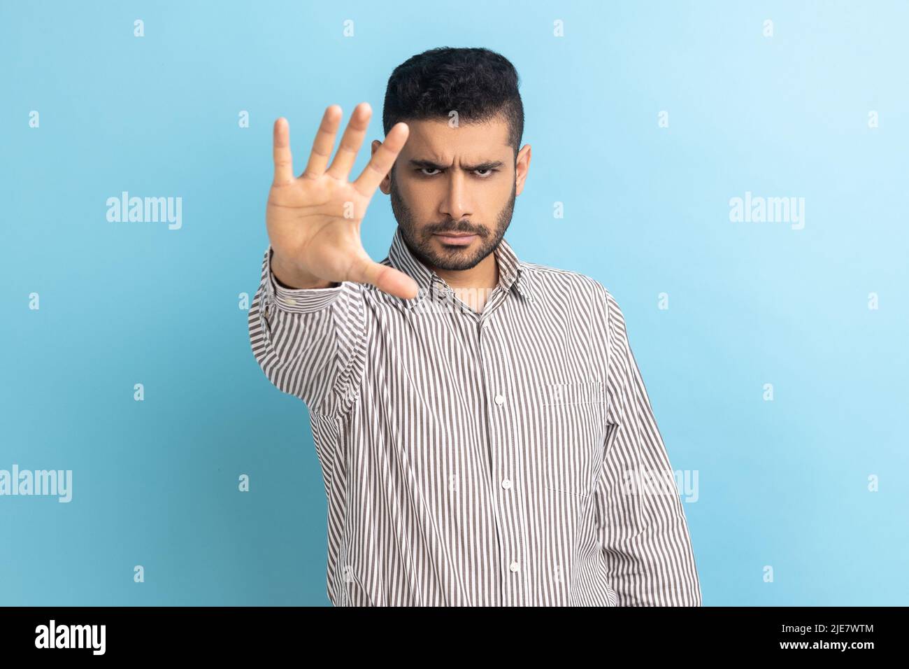Ernsthafter Geschäftsmann mit Bartstoppbewegung, die Handfläche zeigt, Warnung vor Gefahren durch Konfliktverbote, Mobbing stoppen, gestreiftes Hemd tragen. Innenaufnahme des Studios isoliert auf blauem Hintergrund Stockfoto