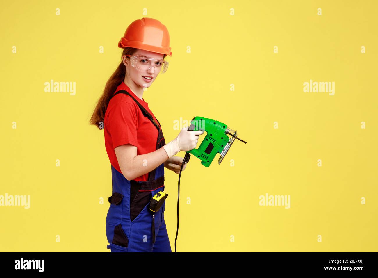 Seitenansicht Porträt einer Bauherrin, die mit der Laubsäge arbeitet, die Kamera mit ernstem Selbstbewusstsein anschaut, Overalls und Schutzhelm trägt. Innenaufnahme des Studios isoliert auf gelbem Hintergrund. Stockfoto