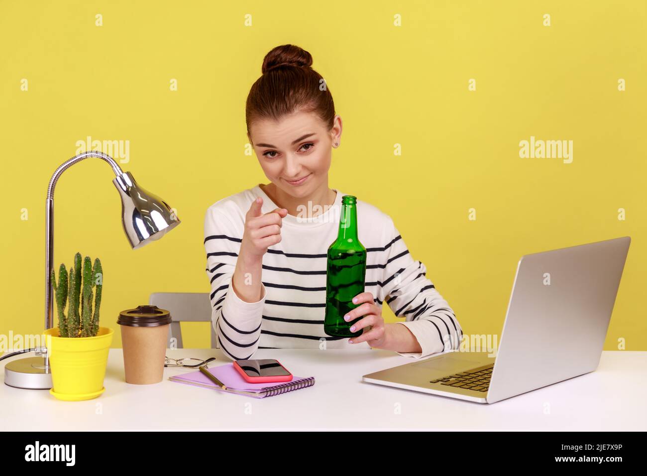 Porträt einer attraktiven jungen erwachsenen Frau im Büro, die eine Bierflasche in den Händen hält und mit einem Lächeln auf die Kamera zeigt. Studio-Innenaufnahme isoliert auf gelbem Hintergrund. Stockfoto