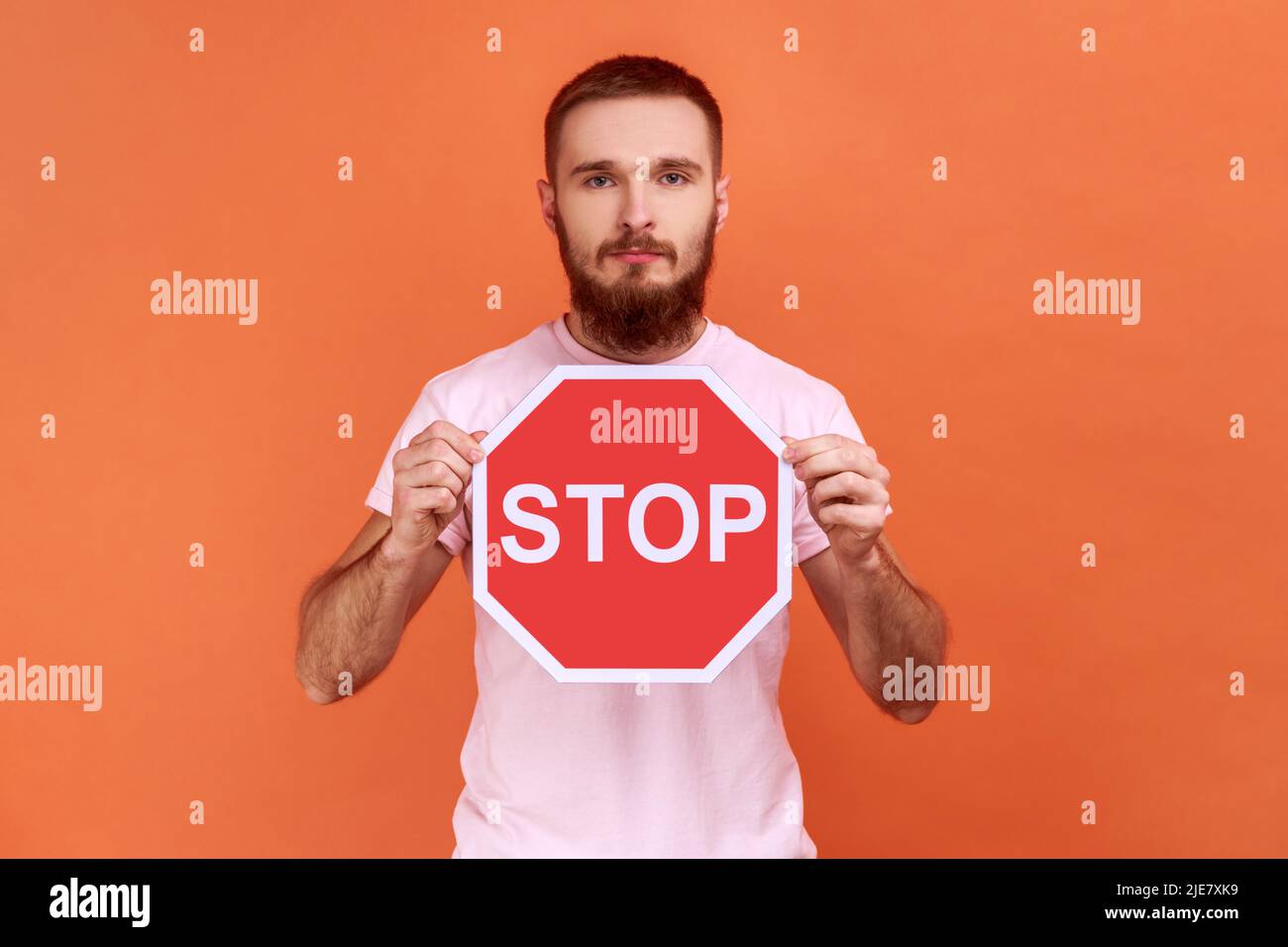 Porträt eines seriös beherrten bärtigen Mannes mit rotem Stoppschild, der mit ernstem Ausdruck die Kamera anschaut und ein pinkfarbenes T-Shirt trägt. Innenaufnahme des Studios isoliert auf orangefarbenem Hintergrund. Stockfoto