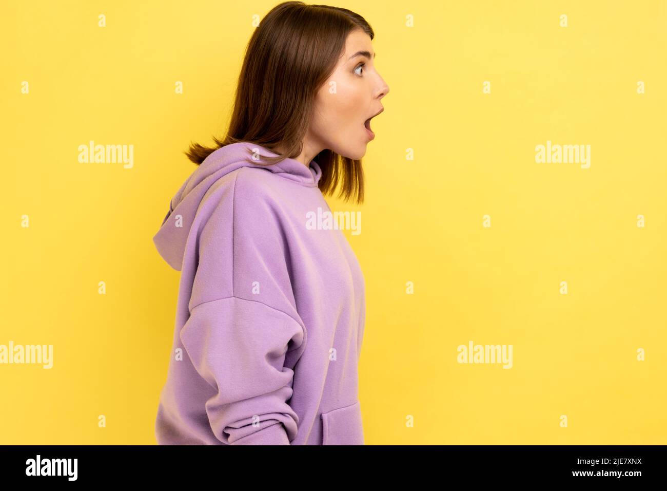 Profil der überraschten dunkelhaarigen Frau, die mit schockierten Gesichtsausdruck zur Seite schaut, hält den Mund weit offen und trägt einen violetten Hoodie. Innenaufnahme des Studios isoliert auf gelbem Hintergrund. Stockfoto
