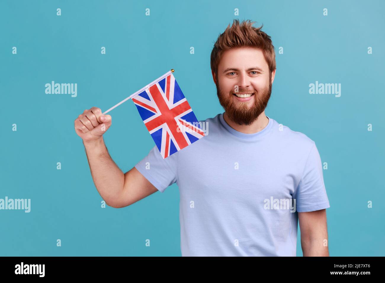 Porträt eines gutaussehenden, zufriedenen bärtigen Mannes, der die Flagge einer konstituierenden Einheit des Vereinigten Königreichs hält und den Tag der britischen Unabhängigkeit feiert. Innenaufnahme des Studios isoliert auf blauem Hintergrund. Stockfoto
