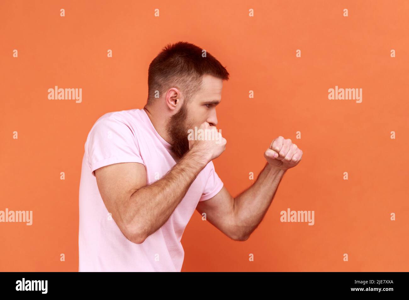 Seitenansicht des bärtigen, aggressiven Kämpfers, der geballte Fäuste bereit zum Boxen hält, Kampfkunsttrainer, Selbstverteidigung, mit pinkem T-Shirt. Innenaufnahme des Studios isoliert auf orangefarbenem Hintergrund. Stockfoto