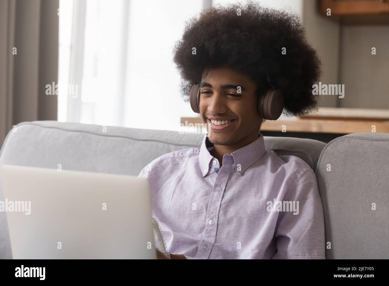 Ein fröhlicher afrikanischer Mann mit Kopfhörern sitzt mit einem Laptop auf dem Sofa Stockfoto