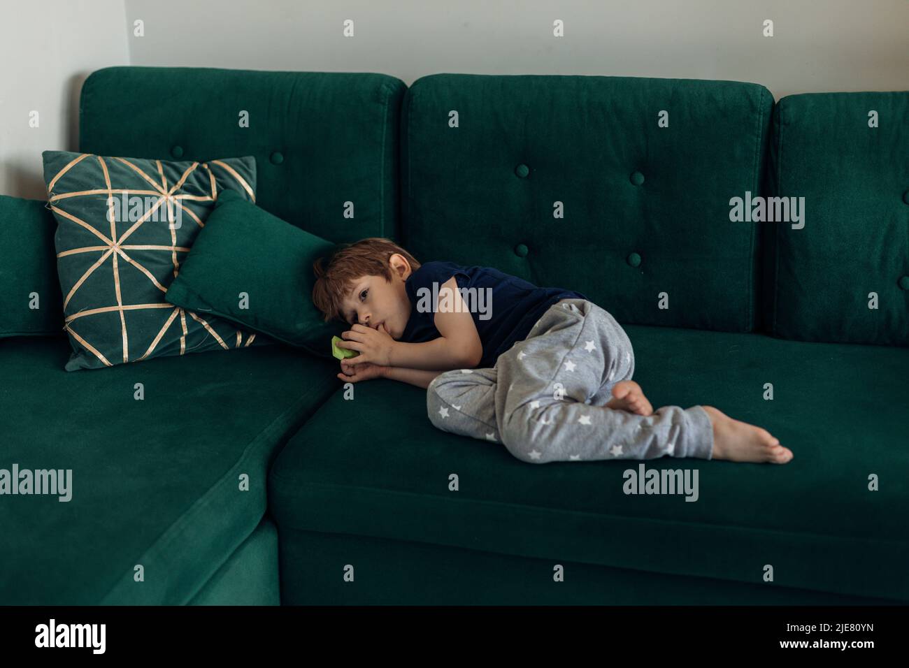 Kleiner Junge in Schlafbekleidung liegt auf einem großen grünen modernen Sofa im Wohnzimmer und saugt Finger. Schlechtes Habbit und Benehmen von kleinen Kindern. Ziehen Sie die Finger hinein Stockfoto