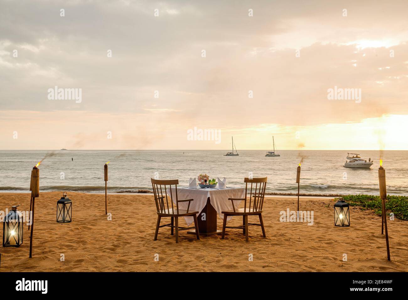 Romantisches Abendessen bei Sonnenuntergang am Strand. Tisch-Flitterwochen-Set für zwei Personen mit luxuriösem Essen, Gläsern Champagner-Getränken im Restaurant mit Meerblick und Yachten Stockfoto