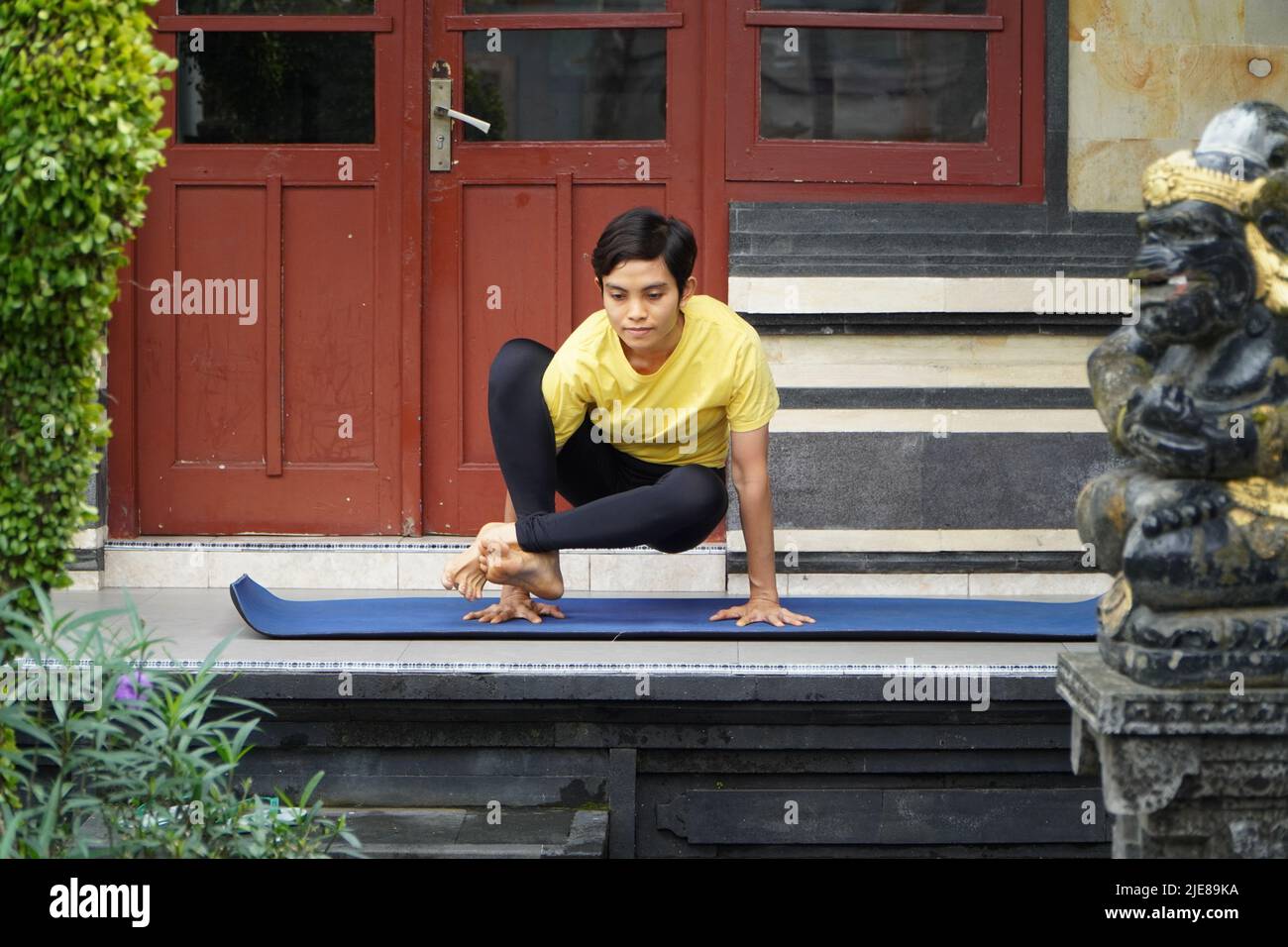 Auf seiner Terrasse übt ein junges asiatisches Mädchen mit einem atemberaubenden Aussehen Yoga, während es einen kurzen Haarschnitt, ein gelbes Hemd und schwarze Leggings trägt. S Stockfoto
