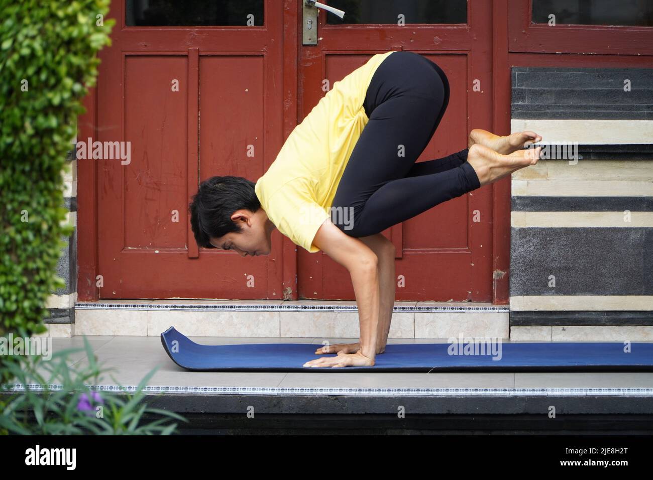 Auf seiner Terrasse übt ein junges asiatisches Mädchen mit einem atemberaubenden Aussehen Yoga, während es einen kurzen Haarschnitt, ein gelbes Hemd und schwarze Leggings trägt. S Stockfoto