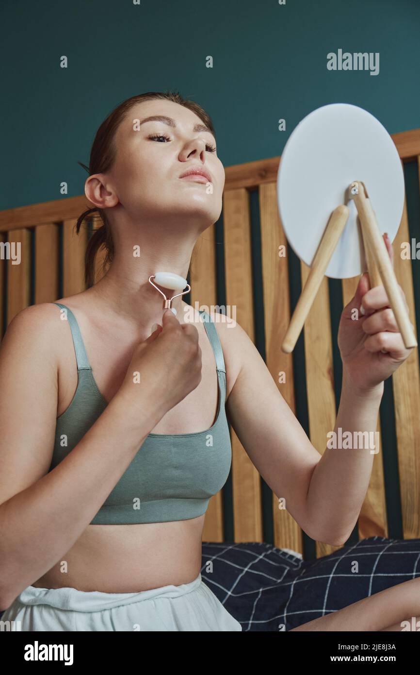 Junge Frau mit Jade Gesichtswalze für Gesichtsmassage sitzen auf dem Bett im Schlafzimmer Blick in den Spiegel Stockfoto