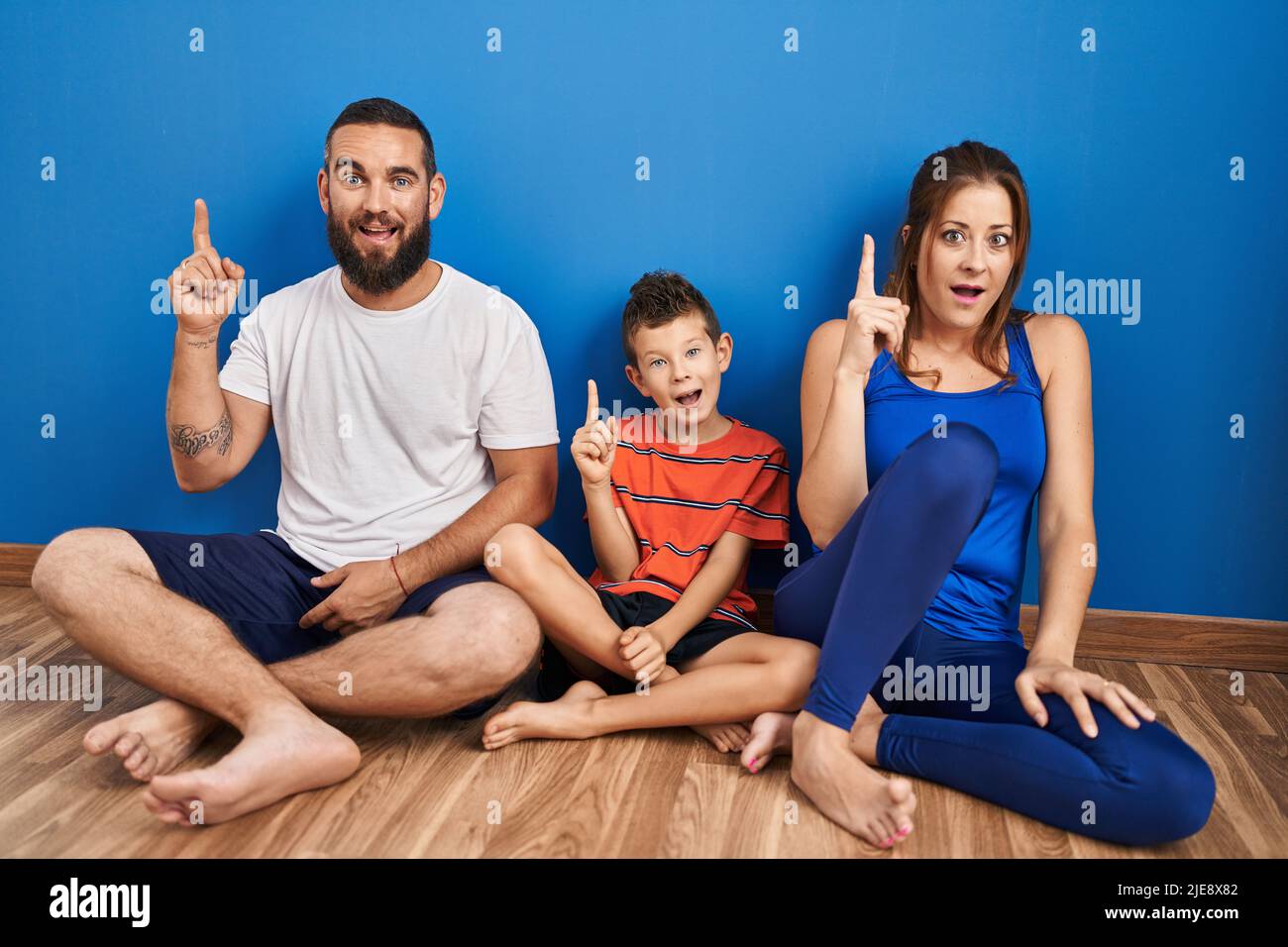 Dreiköpfige Familie, die zu Hause auf dem Boden sitzt und mit dem Finger nach oben zeigt, mit einer erfolgreichen Idee. Begeistert und glücklich. Stockfoto