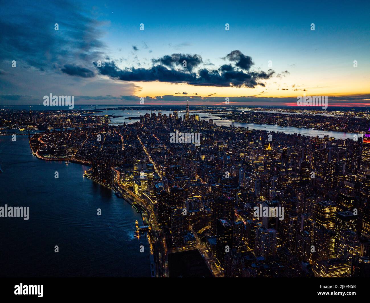 Atemberaubende Panoramaaufnahme der Stadt, die vom Fluss umgeben ist. Beleuchtete Fenster und farbenfroher Sonnenuntergang. Manhattan, New York City, USA Stockfoto