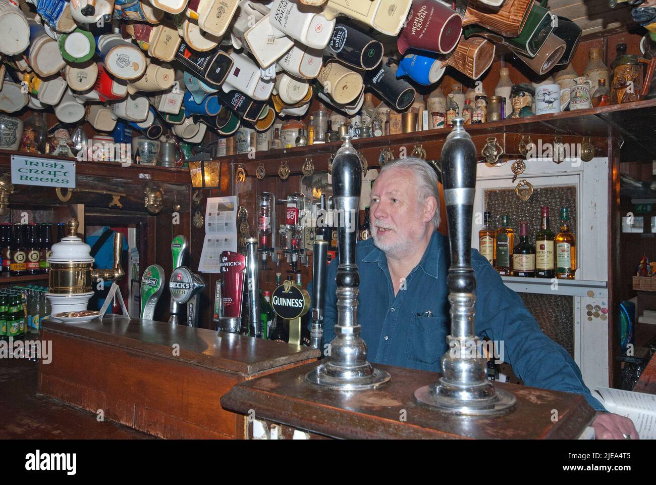 Vermieter hinter der Theke in Nancy's Bar mit Tassen und Krügen, die an der Decke hängen, Ardara, County Donegal, Irland Stockfoto