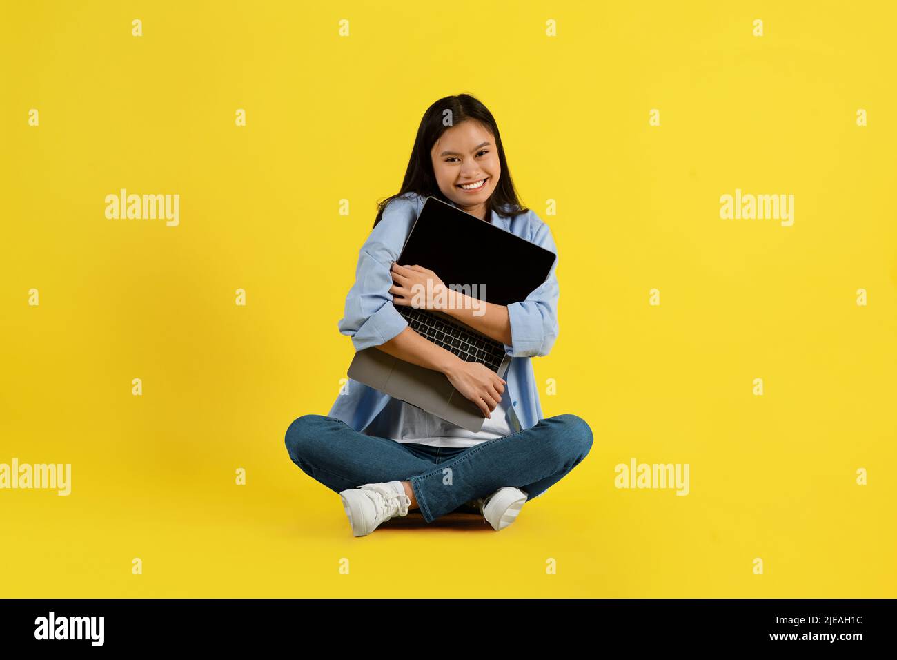 Fröhliche, fröhliche junge asiatische Studentin mit Down-Syndrom sitzt auf dem Boden und umarmt den Laptop Stockfoto