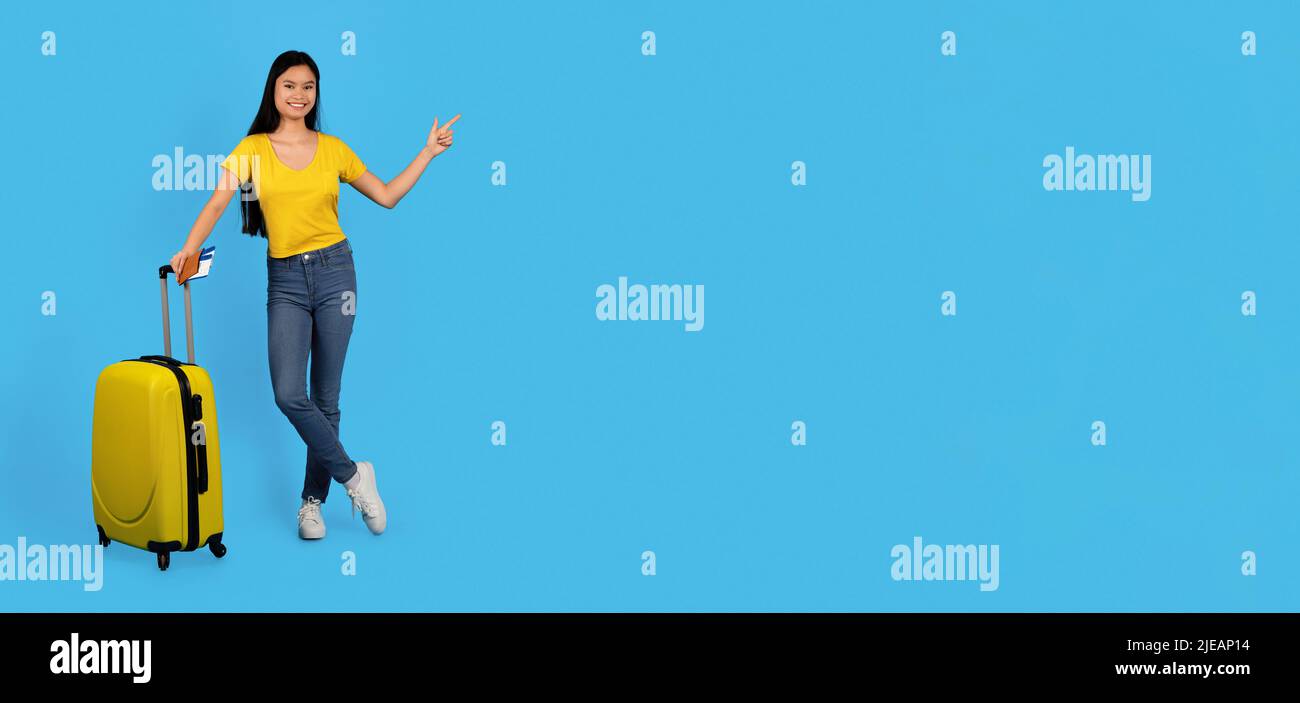 Fröhliche, tausendjährige japanische Hündin in gelbem T-Shirt mit dem Koffer, der mit dem Finger auf den leeren Raum zeigt Stockfoto