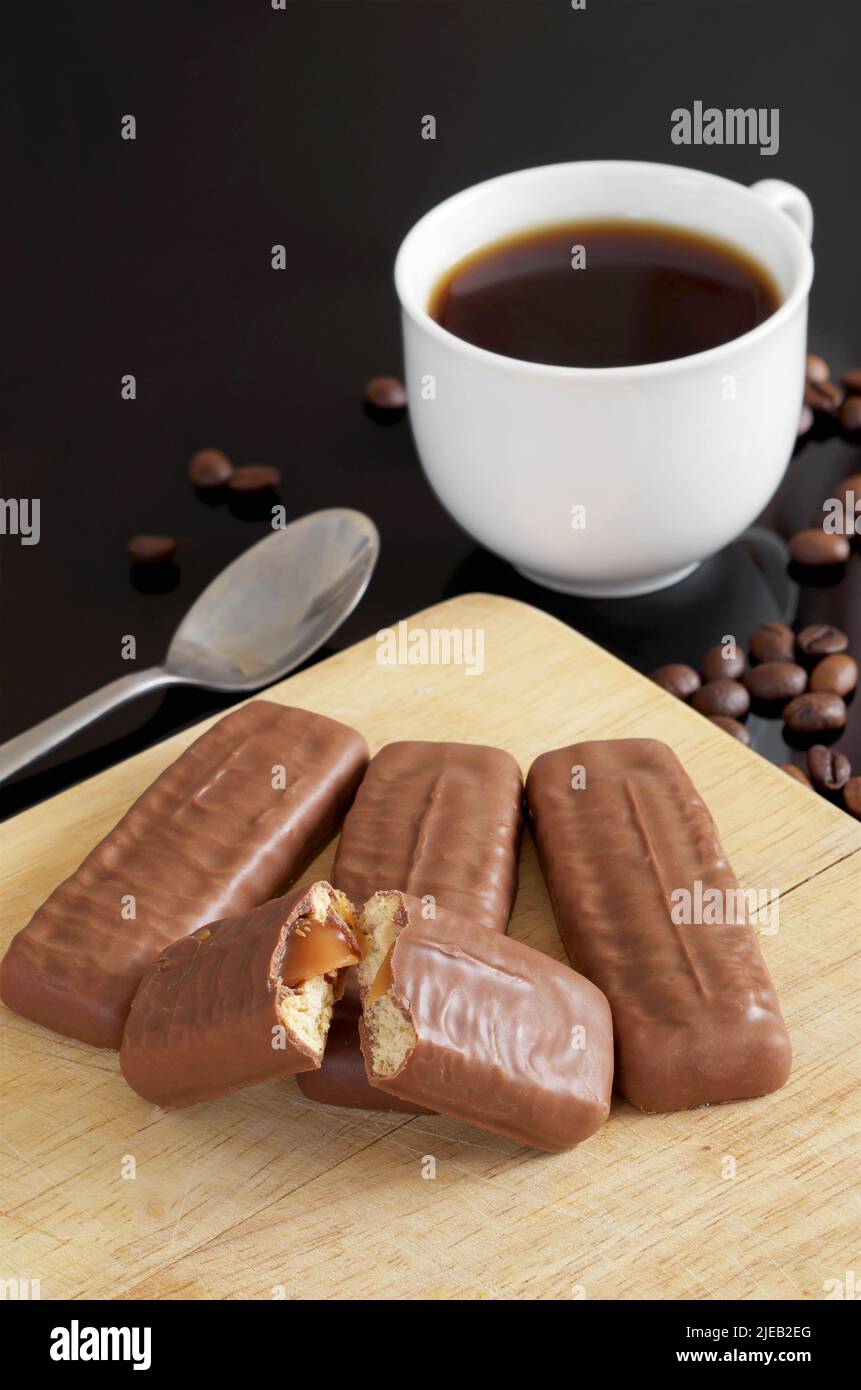 Shortcake-Keksbar mit Karamell und voll mit Milchschokolade überzogen auf Schneidebrett und Tasse Kaffee auf schwarzem Tisch Stockfoto