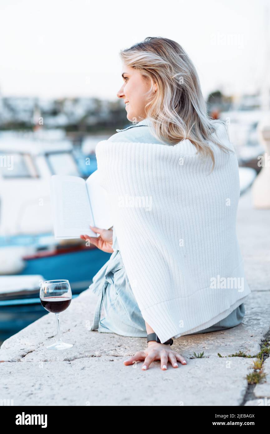 Eine junge Frau mit welligen, lockeren Haaren sitzt mit einem Buch in den Händen auf dem Pier, neben ihr steht ein Glas Rotwein. Stockfoto