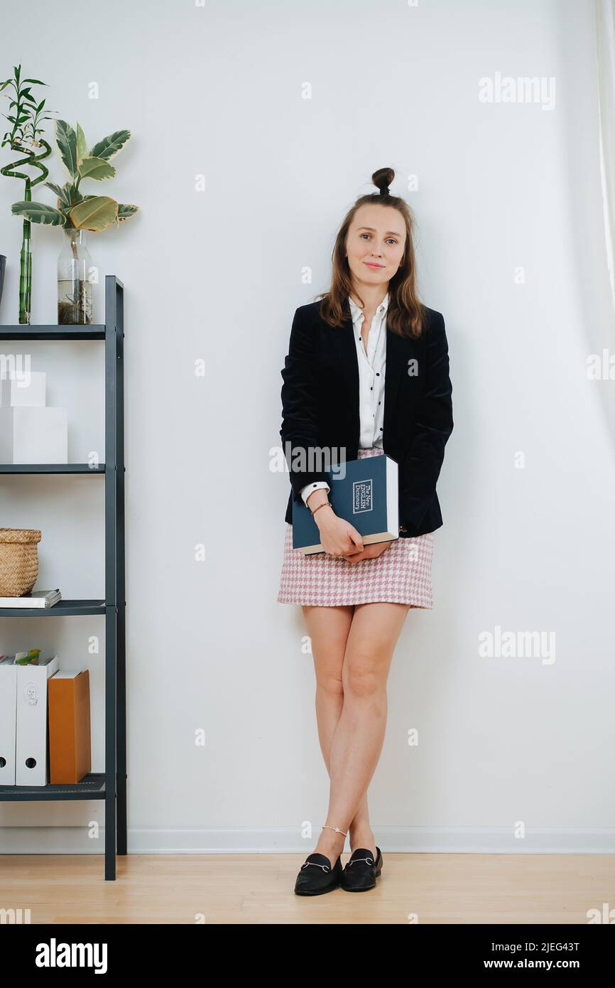 Eine positive Frau, die sich zu Hause an der Wand lehnt und die Kamera anschaut. Sie hat ihre Haare in einem Knoten, trägt einen kurzen karierten Rock und eine Jacke. Stockfoto