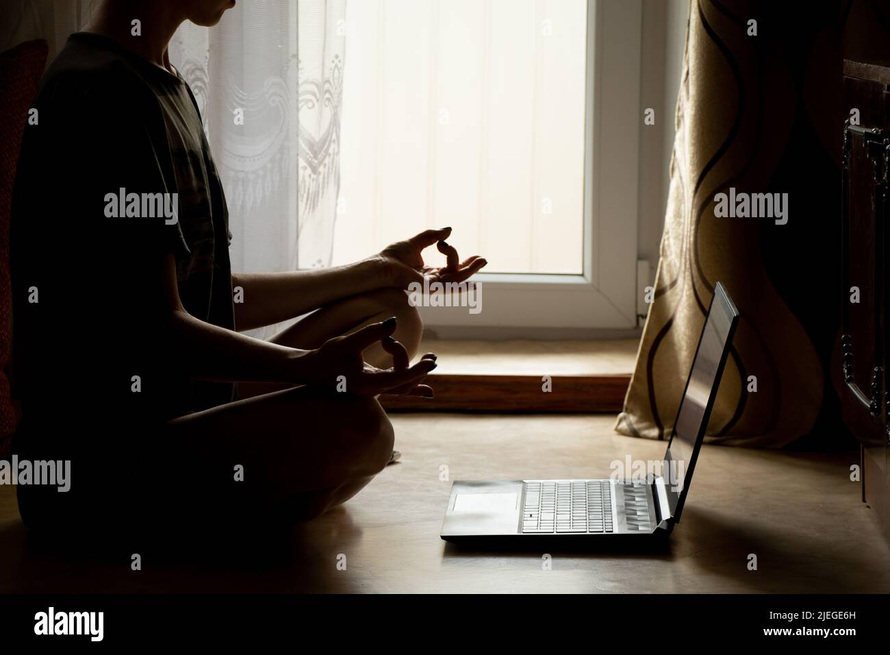 Ein Mädchen macht Yoga in der Nähe eines Laptops auf dem Boden eines Hauses in der Nähe eines Fensters im Dunkeln, einer Online-Videostunde über Meditation, Yoga und Miditatsi Stockfoto