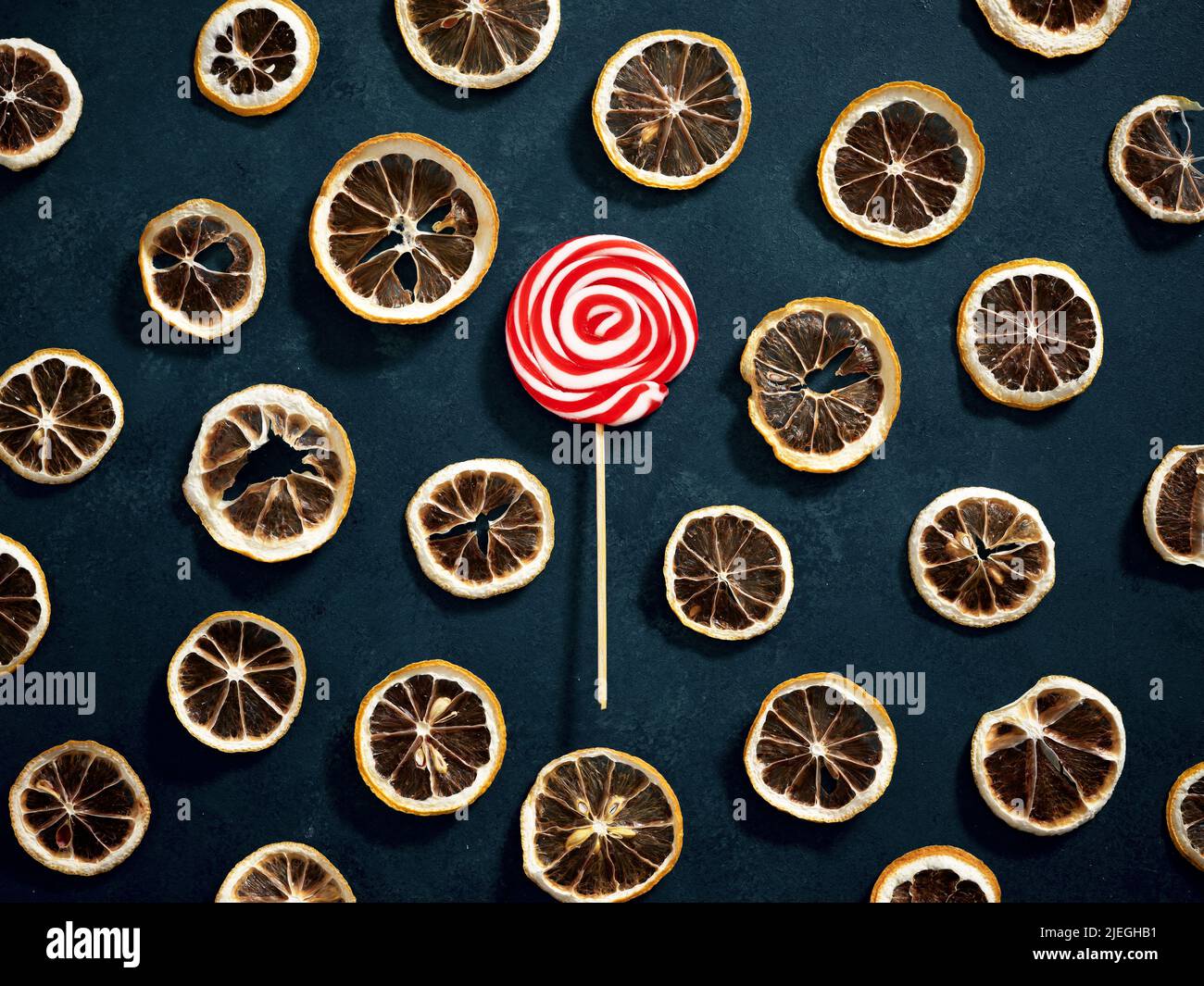 Rot und weiß gefärbter, runder Candy-Lollipop mit trockenen Zitronenscheiben auf blauem strukturiertem Hintergrund. Süßes und saures Essen im Kontrast. Stockfoto