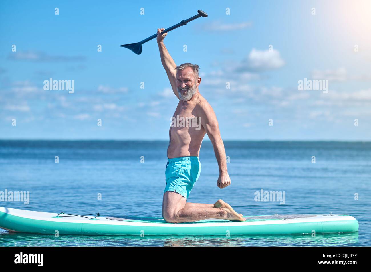 Mann, der das Paddel hebt, lächelt und die Kamera auf dem Surfbrett kniet Stockfoto