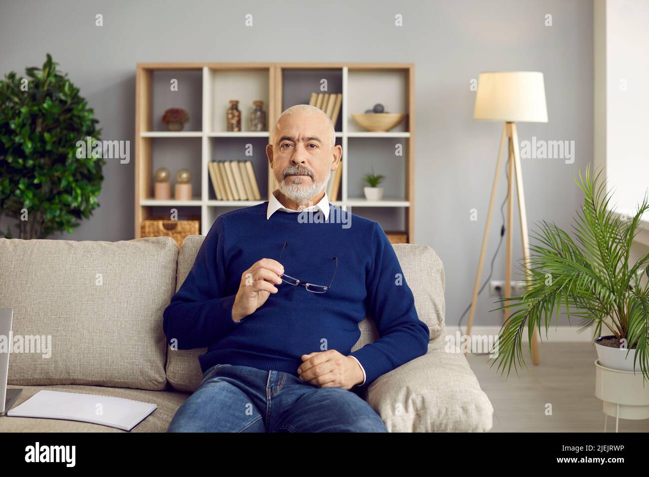 Porträt eines hübschen und stilvollen unrasierten kahlen älteren Mannes, der im Heimbüro auf dem Sofa sitzt. Stockfoto