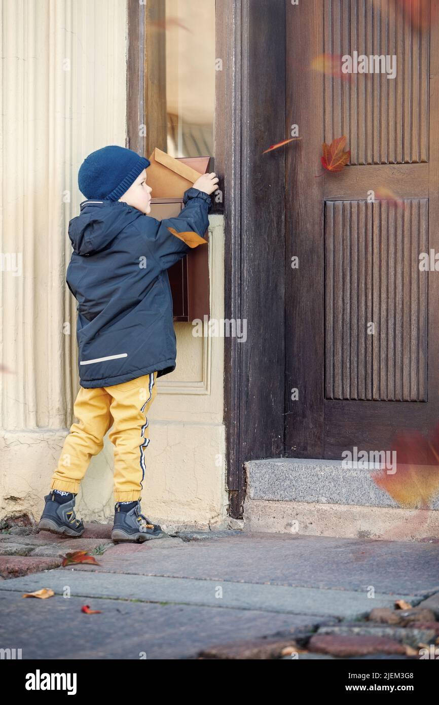 Netter Junge, der Post mit Briefkasten in der Nähe des Hauses sendet oder empfängt. Im Herbst bläst der Wind bunte Baumblätter. Stockfoto