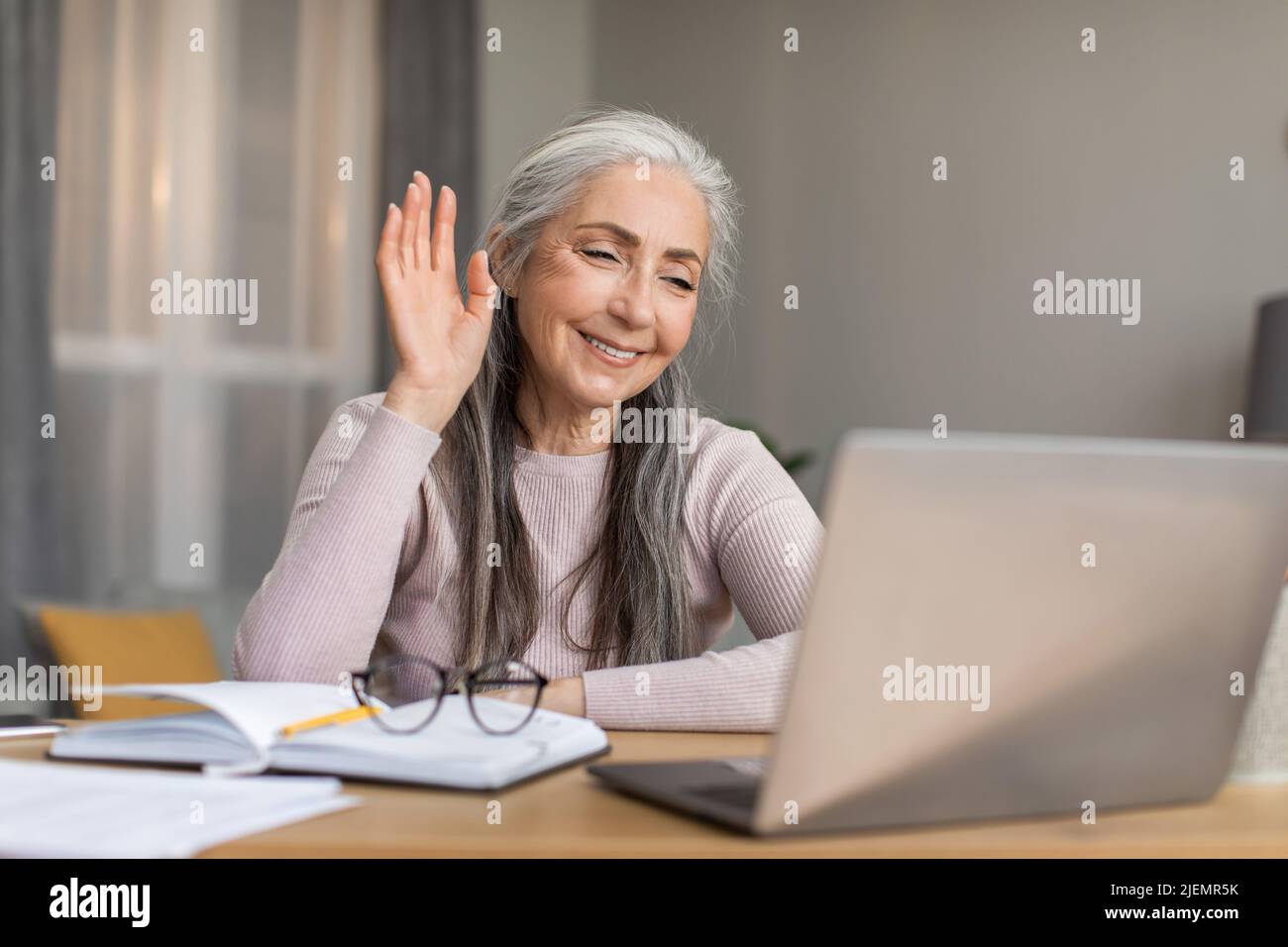 Eine fröhlich lächelnde europäische ältere Frau mit grauem Haar, die ihre Hand am Computer winkte, kam aus der Ferne zu einem Treffen Stockfoto