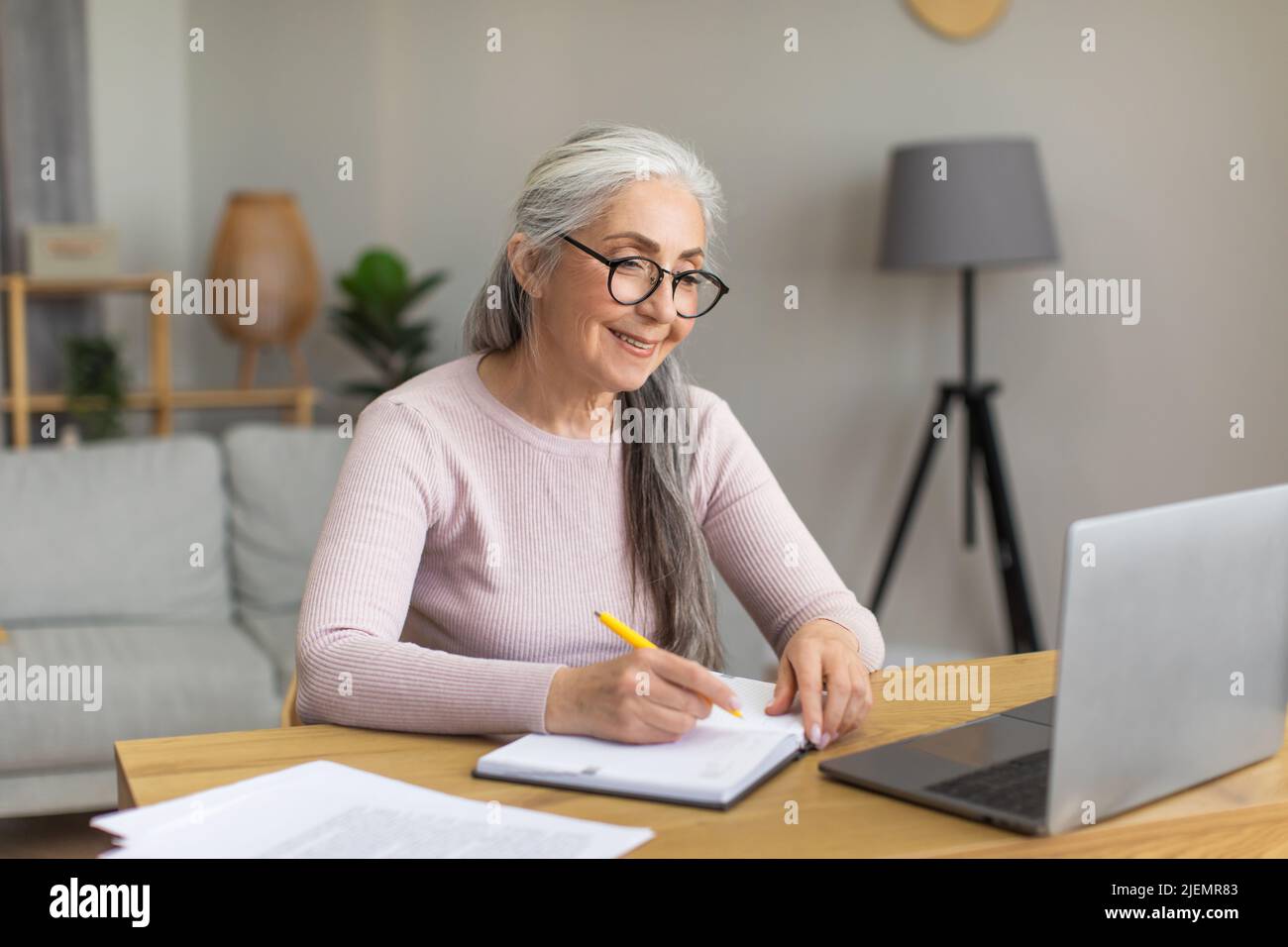 Lächelnde europäische alte Frau mit grauen Haaren in einer Brille schaut auf den Computer, macht sich Notizen am Tisch Stockfoto
