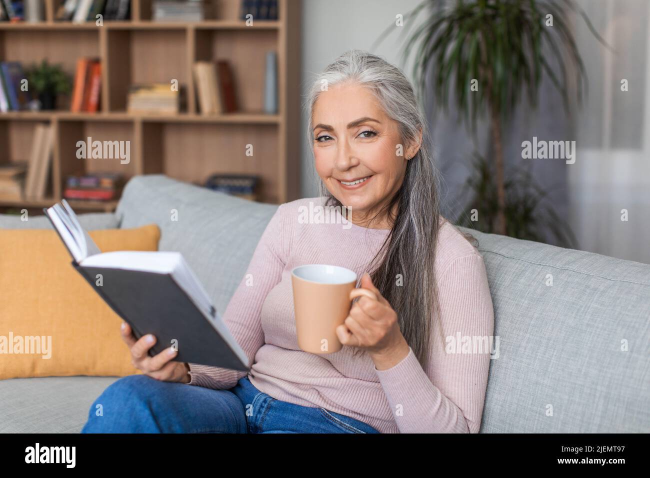 Fröhliche ältere Frau aus europa mit grauen Haaren trinkt Kaffee und liest in der Freizeit Lieblingsbuch Stockfoto