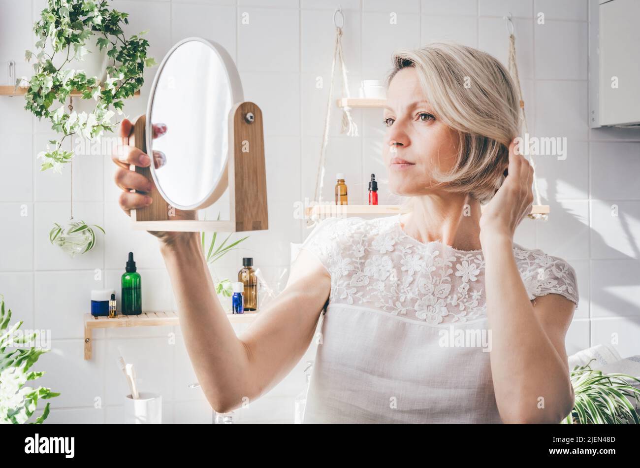 Frau mittleren Alters, die im hellen Badezimmer im Spiegel mit biophilem minimalistischem Design schaut. Pflege der alternden Haut. Gut Stockfoto