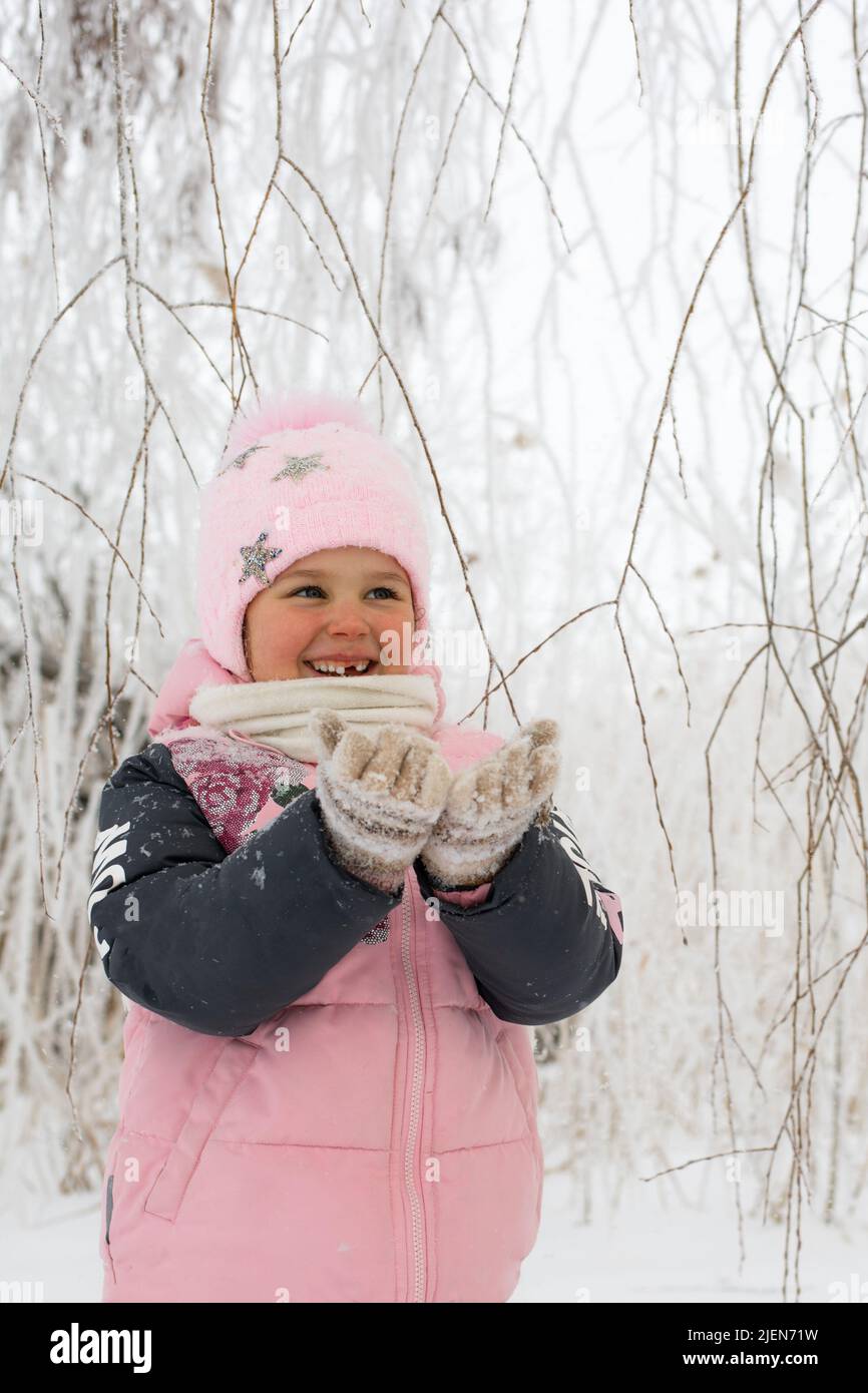 Kleines glückseliges Mädchen mit roten Wangen und einem breiten Lächeln mit fehlenden Vorderzahn, das warme Winterkleidung trägt und Schnee mit verschneiten Bäumen im Hintergrund fängt Stockfoto
