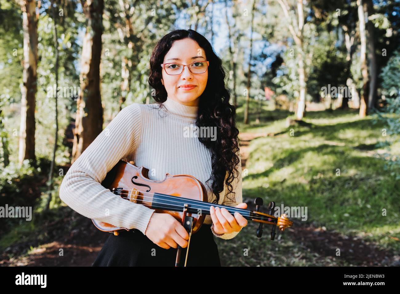 Junge Brünette Frau mit einer Brille, die draußen im Wald eine Geige hält. Stockfoto