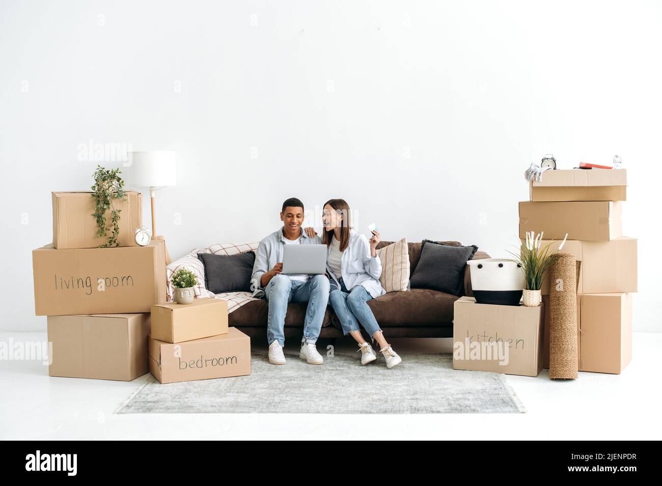 Online-Shopping. Glückliches Paar, ein hispanischer Kerl und ein kaukasisches Mädchen, sitzen in ihrer neuen Wohnung im Wohnzimmer zwischen Kartons mit Dingen, machen Online-Shopping für zu Hause mit Laptop, bestellen Lieferung Stockfoto